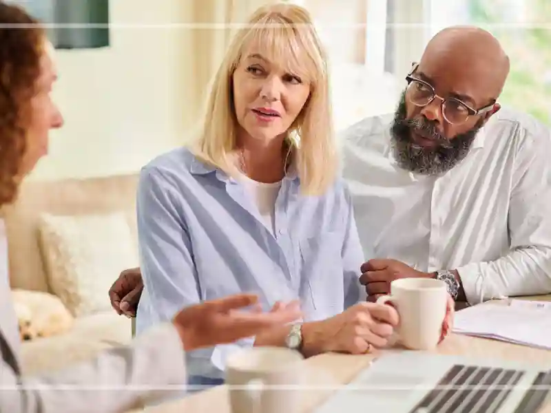 Three people having a discussion at a table with coffee mugs, laptop and documents.